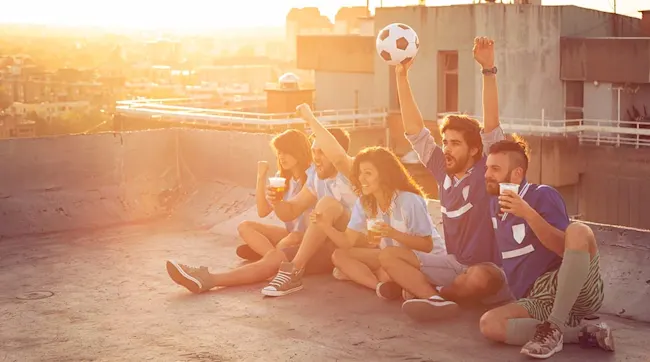 Een groep mensen zit op een dak met een voetbal en drankjes, juichend met de stad op de achtergrond.