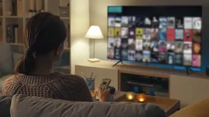 Een vrouw zit op de bank voor een tv met een menuscherm. Een lamp en kaarsen staan op een kast.