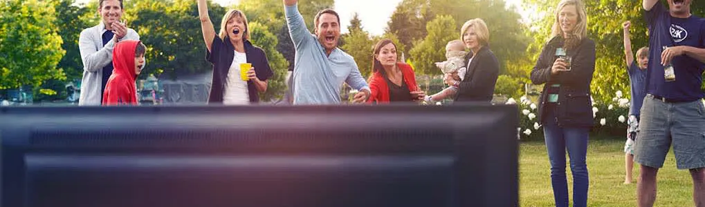 Mensen kijken naar een tv buiten op gras. Ze juichen en houden drankjes vast. Achtergrond: bomen en zon.