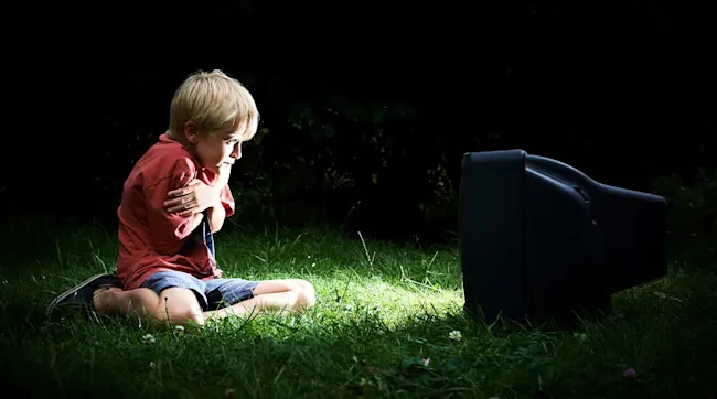 Een jongen zit op gras voor een oude, zwarte televisie in het donker en omarmt zichzelf.