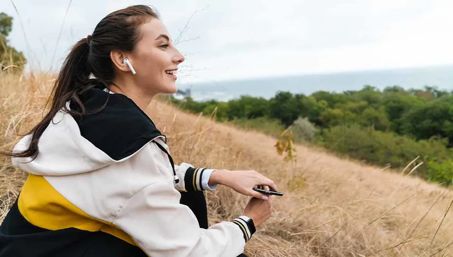 Een vrouw met oortjes en een hoodie zit in hoog gras. Op de achtergrond zijn bomen en de zee te zien.