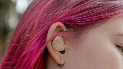 Duidelijke foto van een oor met een beige oortje en een zilveren oorbel, met roze geverfd haar op de achtergrond.