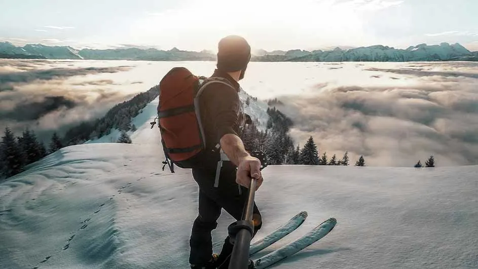Een persoon met een rode rugzak staat op ski's in de sneeuw met bergen en wolken op de achtergrond.
