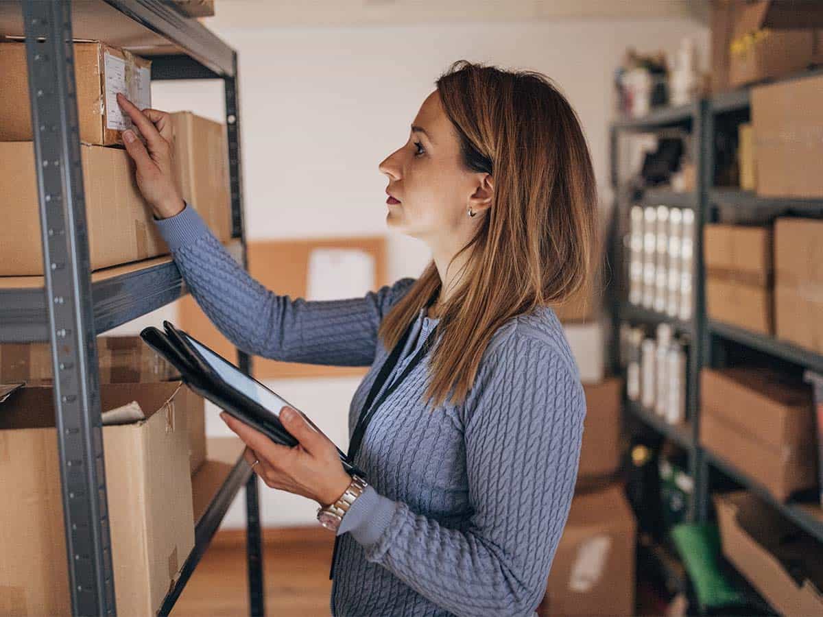 Una donna con i capelli biondi che tiene in mano un dispositivo mobile con uno schermo e controlla l'etichetta di inventario su un pacco