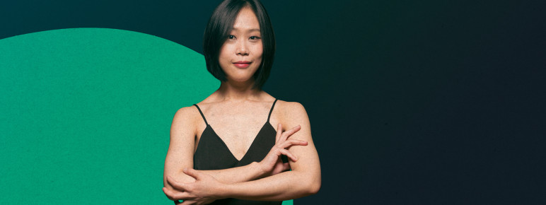 Pianist Yeol Eum Son standing with her arms crossed elegantly, lightly smiling at the camera, against a dark green background with a lighter green circle.