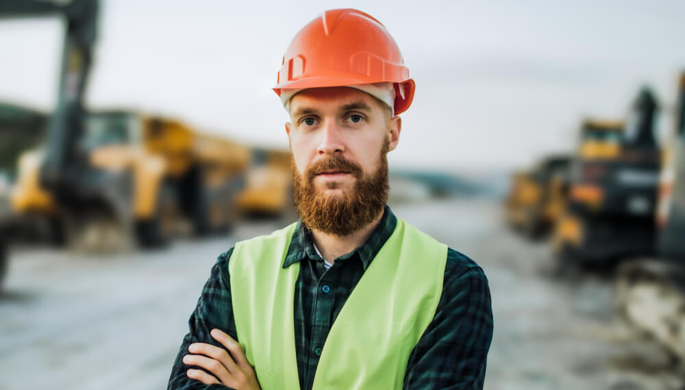 An engineer wearing a safety helmet and reflective vest stands confidently with arms crossed, against the backdrop of a construction site