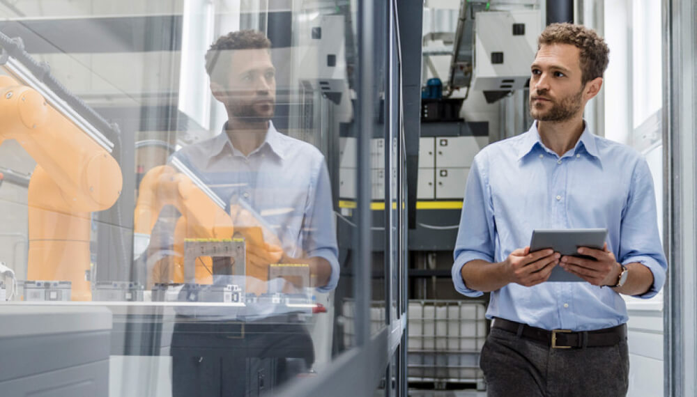 An engineer in a modern factory holding a tablet and inspecting robotic machinery behind a glass panel, reflecting a focus on advanced manufacturing technology