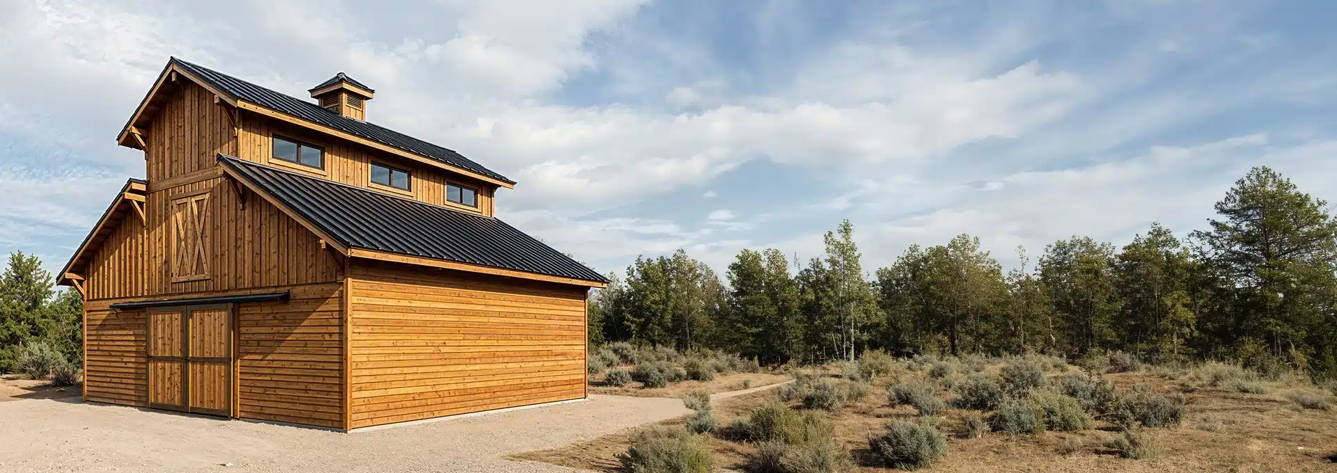 A large wooden barn with a black metal roof stands on a dirt lot surrounded by sparse shrubs and trees under a partly cloudy sky.