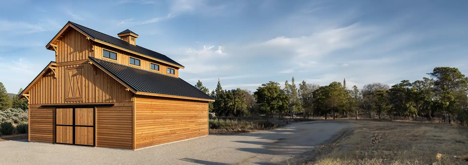 A large wooden barn with a black metal roof stands on a gravel area, bordered by trees and open land under a blue sky.