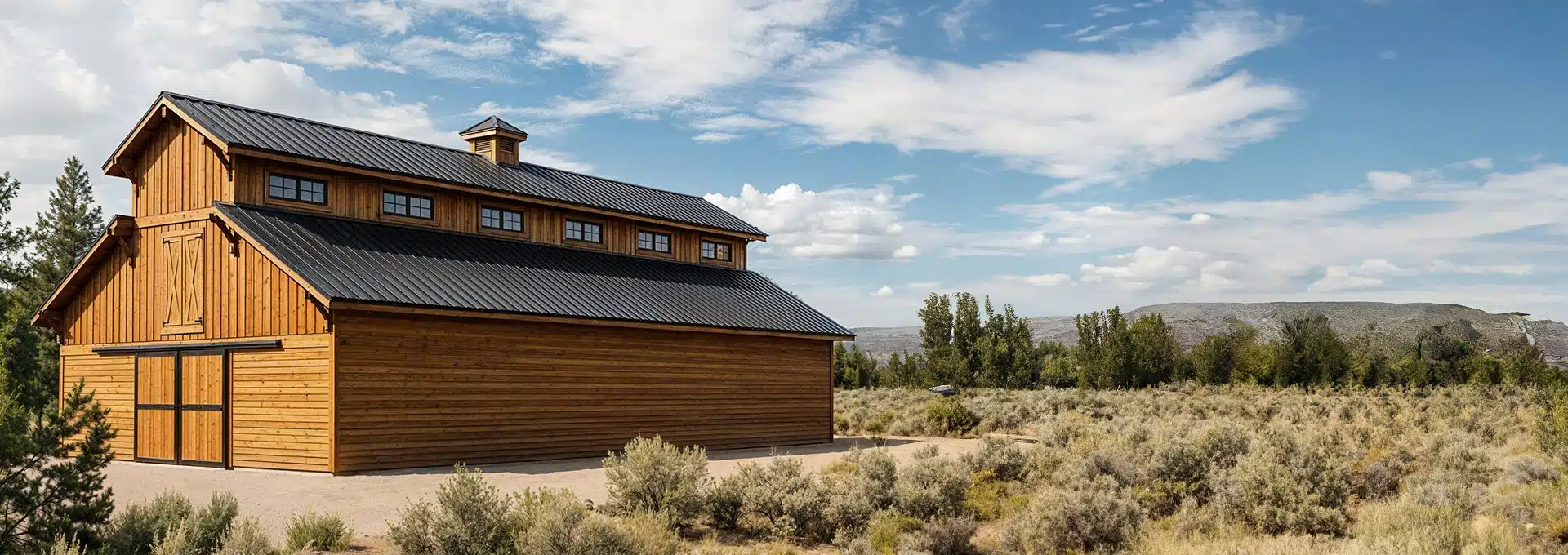 A large wooden barn with a black metal roof stands in a dry, grassy field with shrubs, surrounded by trees and distant hills under a partly cloudy sky.