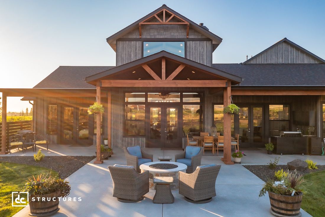 A rustic wooden house exterior with a large patio from our farmhouse kits. The patio features wicker chairs around a fire pit, a dining table, and hanging potted plants. Sunlight casts a warm glow over the scene. A logo for DC Structures appears in the bottom left corner.