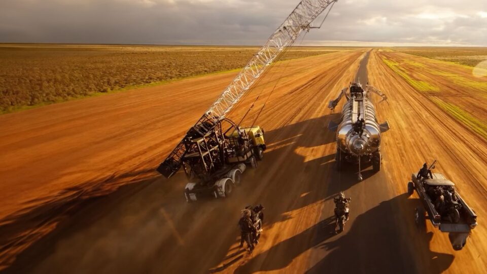 Post-visualization still from Mad Max: Furiosa. A close-up view of a desert chase scene after a disaster. The scene has modified vehicles, including a big tanker truck, a crane-like vehicle, motorbikes, and a pickup truck driving fast across a dusty, reddish-brown road under a dramatic, cloudy sky.