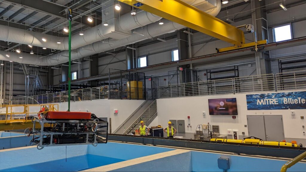 The image shows a rectangular square underwater robot suspended over a large pool of water, and hanging from the roof of a warehouse, with two workers looking on from the side of the pool.