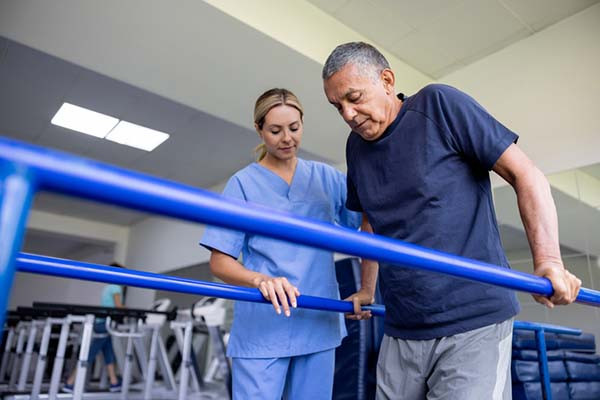 A mature man does physical therapy using balance bars with the assistance of a physical therapist standing by his side.