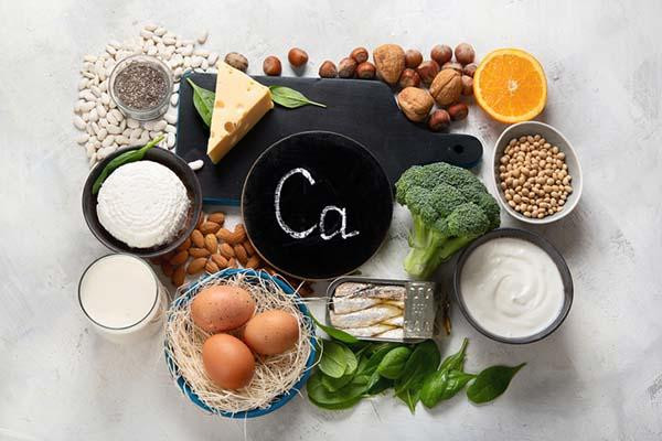 An assortment viewed from above of foods rich in calcium, including cheese, yogurt, milk, eggs, sardines, chickpeas, broccoli, orange, almonds, walnuts, surrounding a black disc with Ca, the chemical symbol for calcium, written on it.