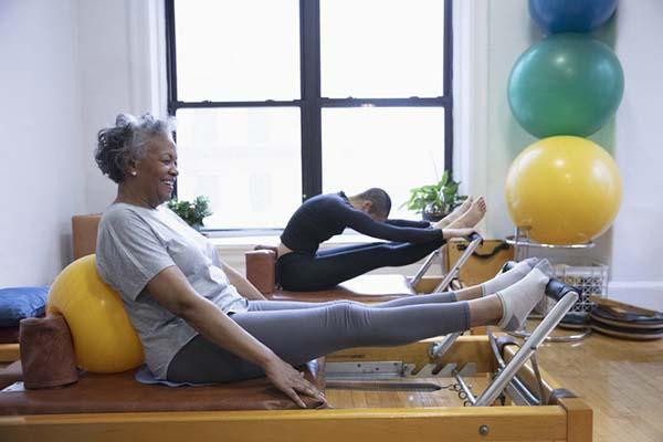 A senior woman smiles while using a Pilates machine in a fitness facility; another person is behind her using the same type of machine, and several large, colorful exercise balls are stacked in a corner.