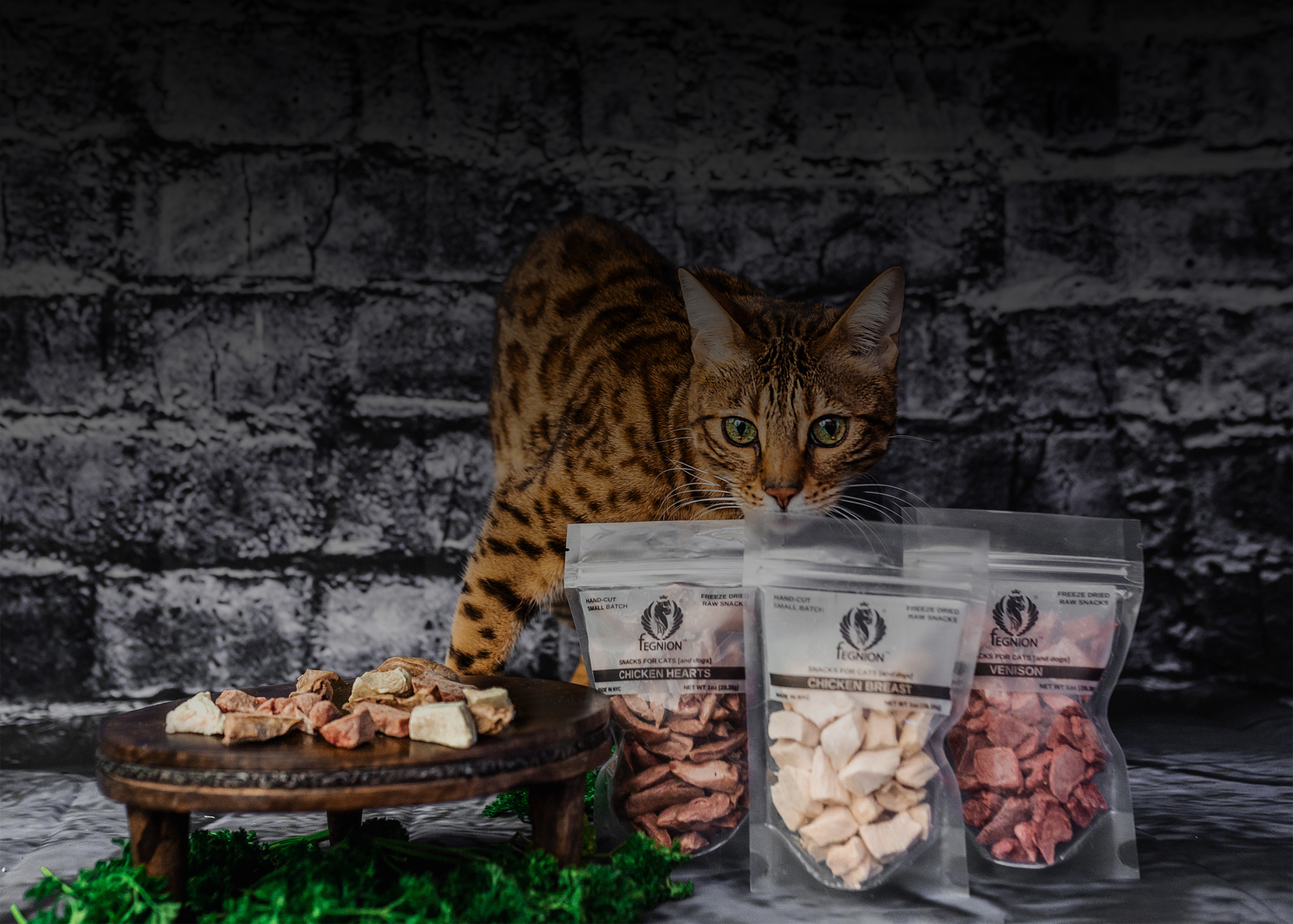 A Bengal cat with green eyes looks over three pouches of Fegnion freeze-dried cat treats: Chicken Hearts, Chicken Breast, and Venison. A wooden tray with loose treats is to the side.