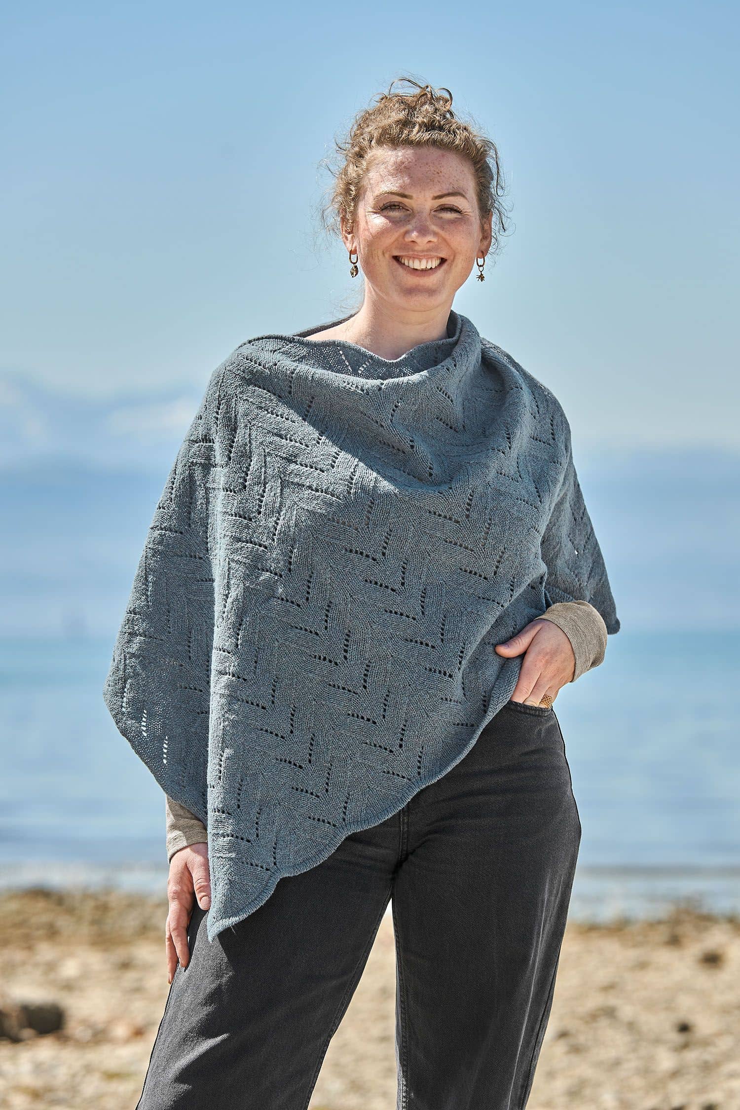 Finkhof-Ajour-Poncho aus reiner Bio-Schurwolle Frau trägt eine schwarze Jeans und einen rauchblauen Poncho mit Ajour Muster vom Finkhof aus bio Merinowolle (kbT)
