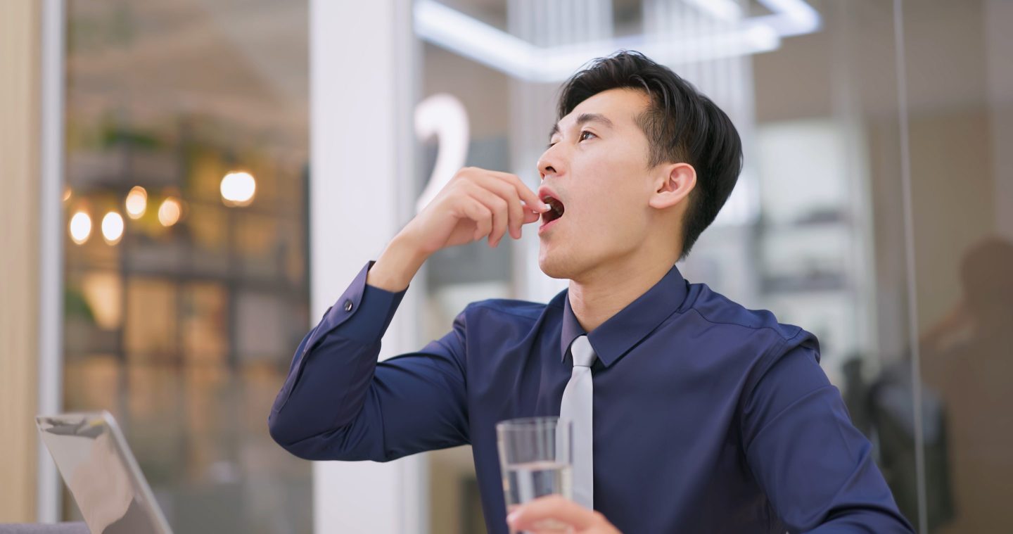 Asian man in shirt and tie taking a pill and holding a glass of water