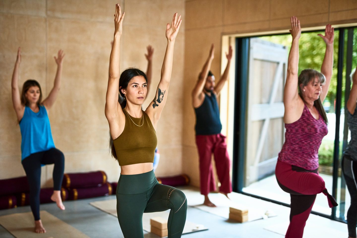 Yoga group standing on one leg with arms above heads, woman in her 30s with tattoo on arm, concentration, focus, balance