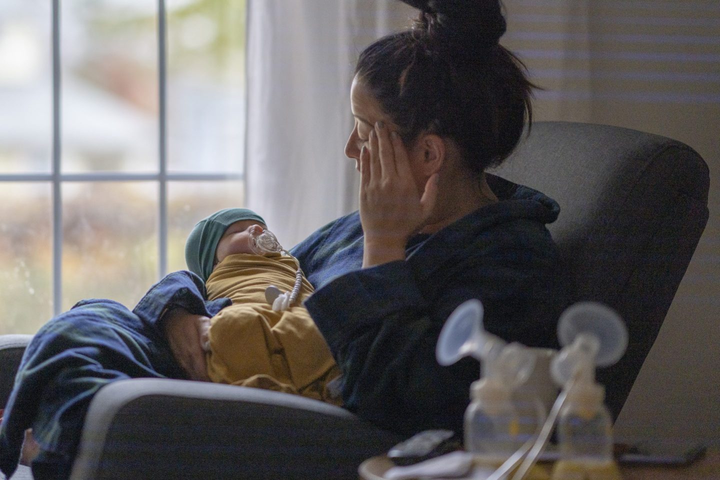 A new mother sitting on a rocking chair in her living room wearing a housecoat holding her newborn baby. There is a breast pump and nursing supplies around.