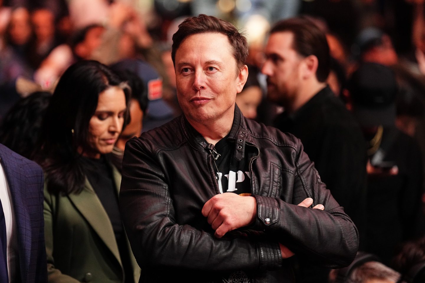 Elon Musk stands with his arms crossed, smirking.