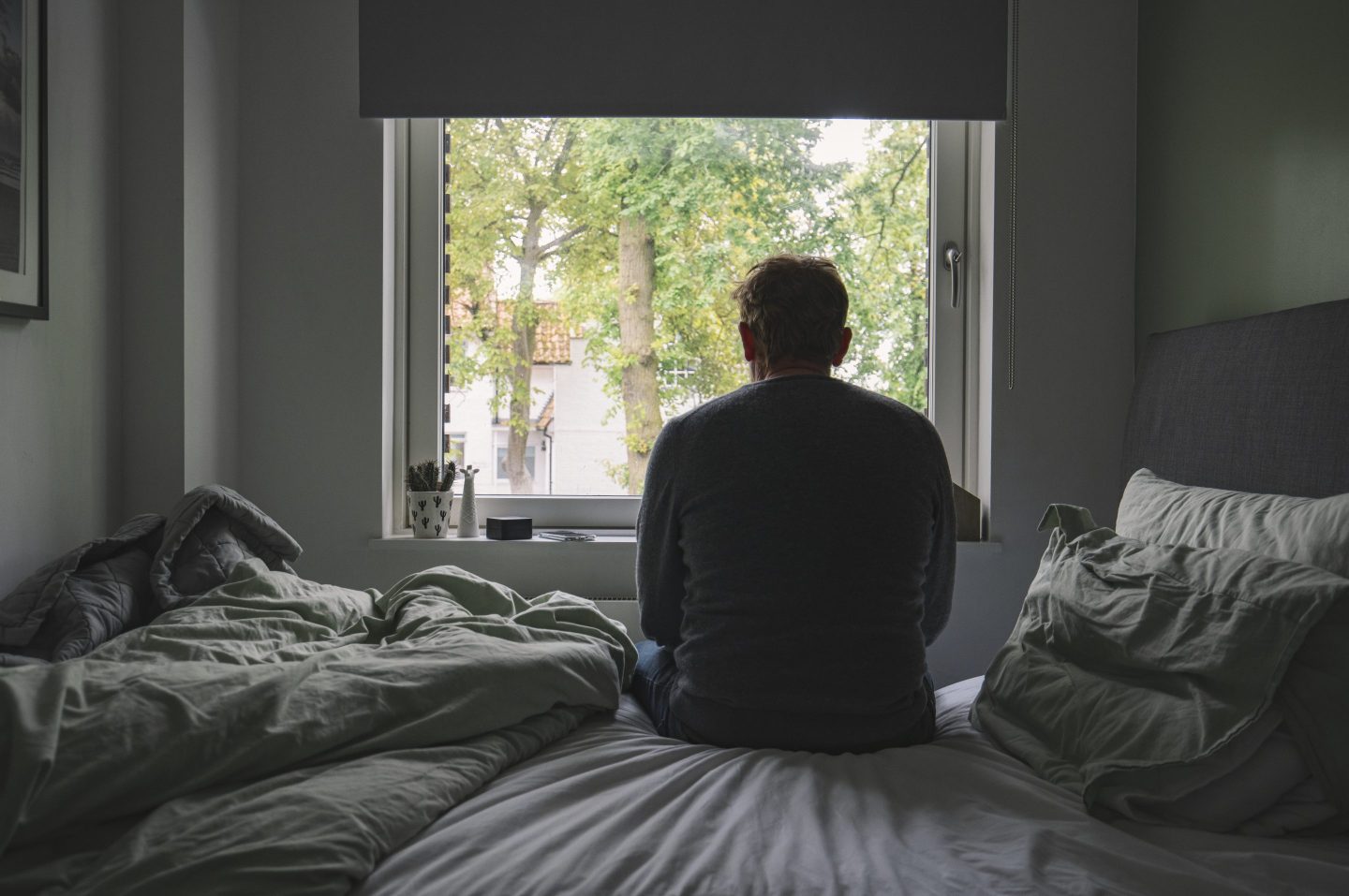 Man, seen from the back, sitting on unmade bed and looking out a window.