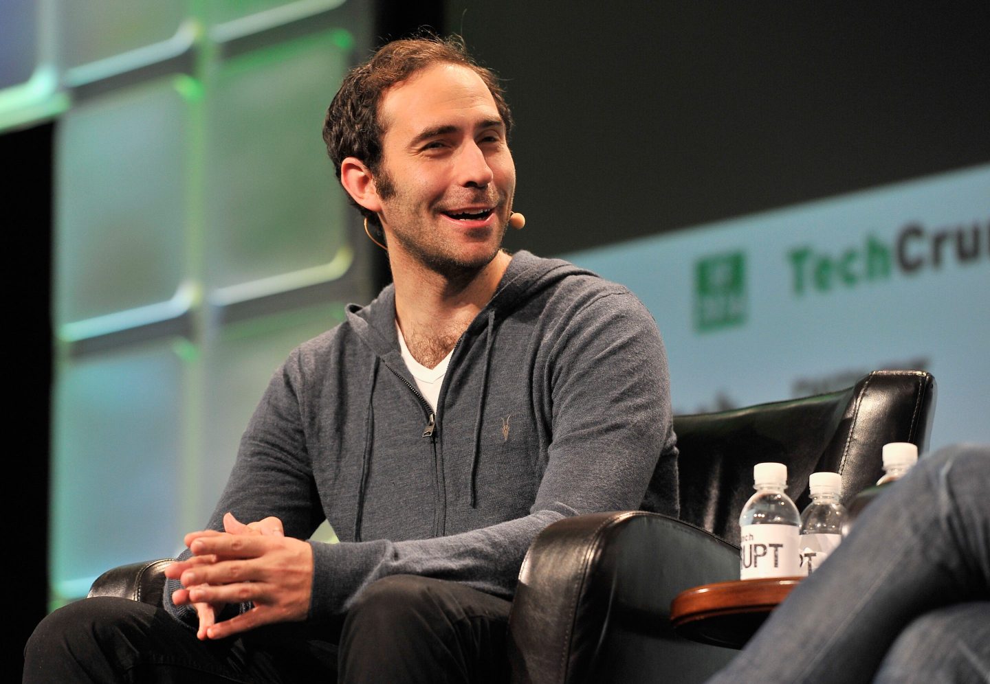 Emmett Shear wears a grey hoodie and speaks onstage to an event.