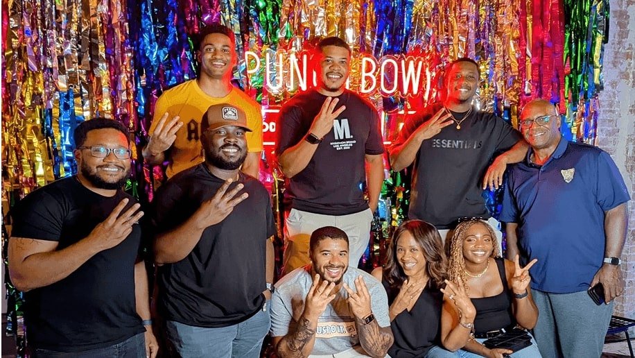 A group of people smiling and posing in front of a colorful backdrop with multicolored foil streamers and a neon sign that reads "PUNCH BOWL," creating a festive and vibrant atmosphere.