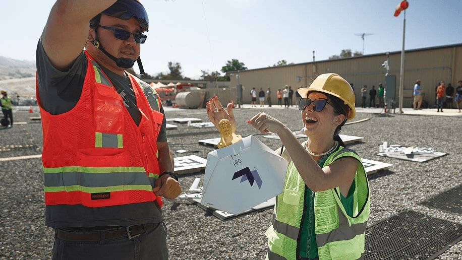 Two people wearing safety vests and helmets celebrate a successful outdoor team-building exercise involving a Wing delivery package, amidst a group of partially visible participants and safety equipment in a gravel-covered area.