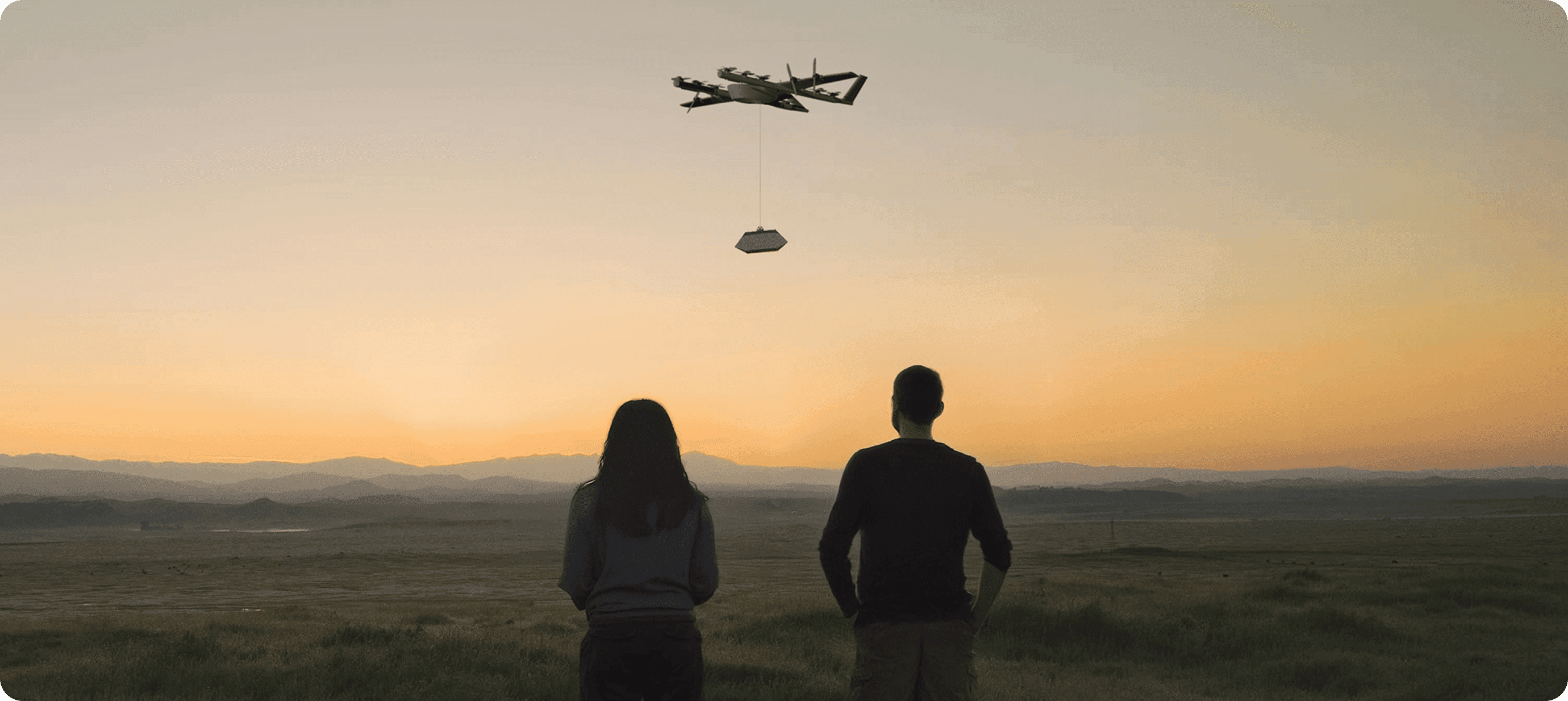 Two silhouetted figures stand facing a sunset, with a package-carrying Wing delivery drone above them.