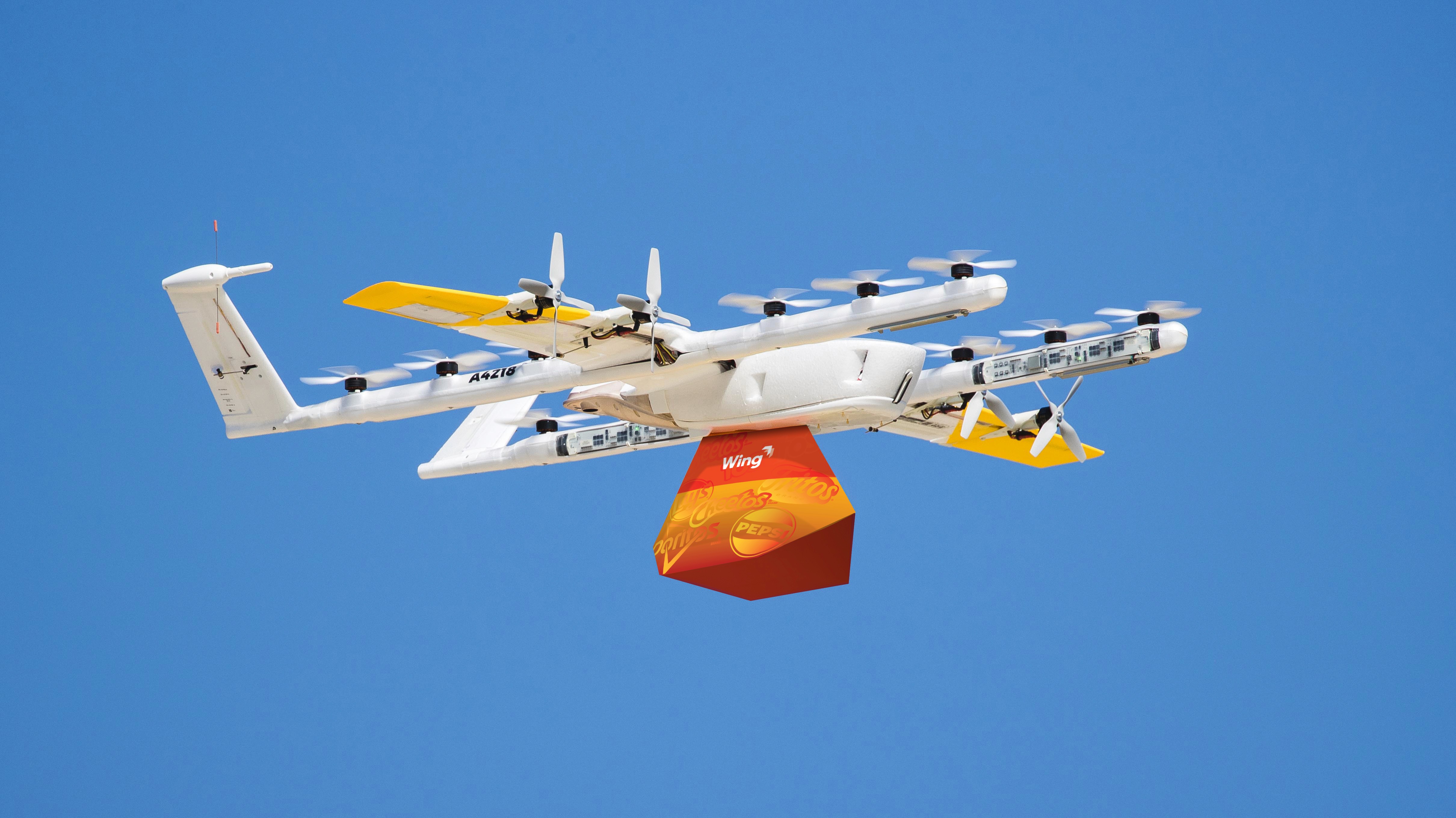 A white and yellow drone in flight against a clear blue sky; it is carrying a red and orange package, demonstrating modern delivery technology.