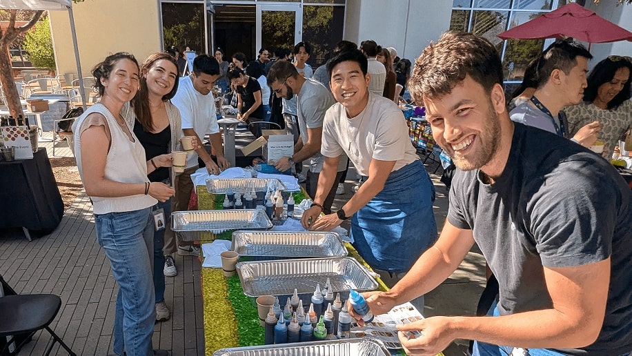 A group of people happily participate in a vibrant outdoor tie-dye activity, surrounded by colorful bottles of dye and large metal trays on tables, under a sunny sky.