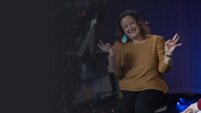 A woman with curly hair and large earrings sits on a stool, smiling and making a hand gesture. She is wearing a mustard-colored top and is positioned in front of a camera setup, with a blurred background.