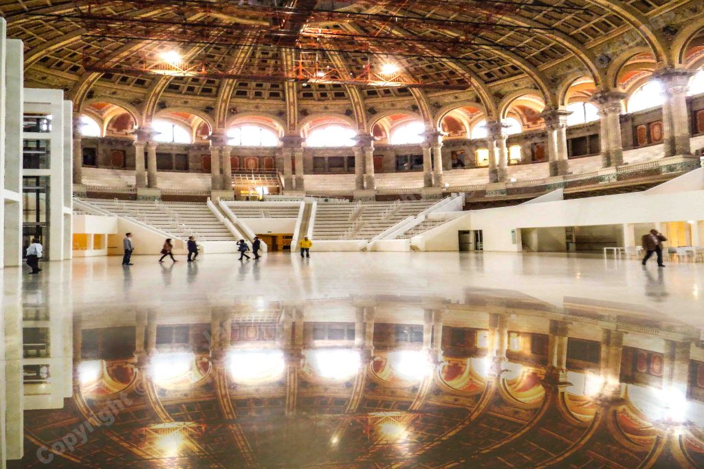 The coloured image depicts the grand entrance and oval hall to the National Museum of Art Barcelona. The vaulted dome with its pillars, arched windows and tiered seating is reflected in the polished marble floor.