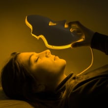 Woman laying down is putting on a face mask with eye, nose, and mouth cutouts. The mask is lit with yellow colored lights from within. 