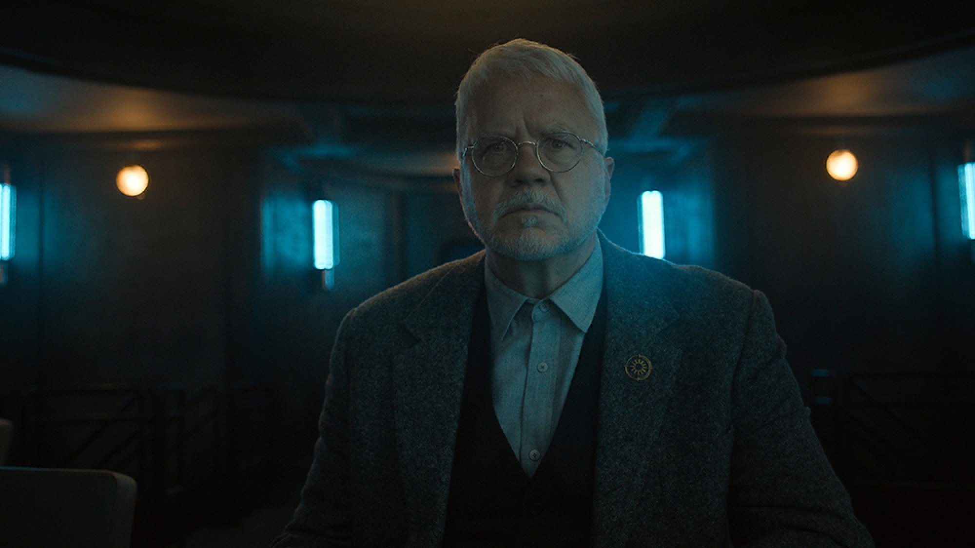 A grey-haired man stands in a dark room lit by blue lights, looking shocked.