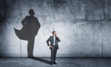 A man in a suit stands against a wall as his shadow shows in the background as if he is wearing a cape like a superhero. 