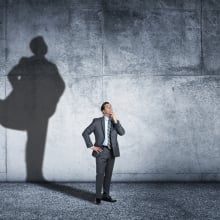 A man in a suit stands against a wall as his shadow shows in the background as if he is wearing a cape like a superhero. 