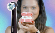 Black text on left side reads "Oral-B iO" against a light blue background. Girl with dark hair holds up a set of fake teeth to the camera.