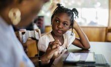 A young girl sits at a table with a tablet device in front of her. She is watching an adult type on the device.