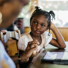 A young girl sits at a table with a tablet device in front of her. She is watching an adult type on the device.