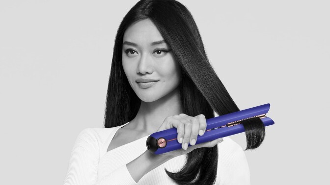 a person uses a blue colored dyson corrale straightener on long, black hair while looking at the camera