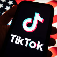 A hand holds out a phone with the TikTok logo in front of an American flag. 