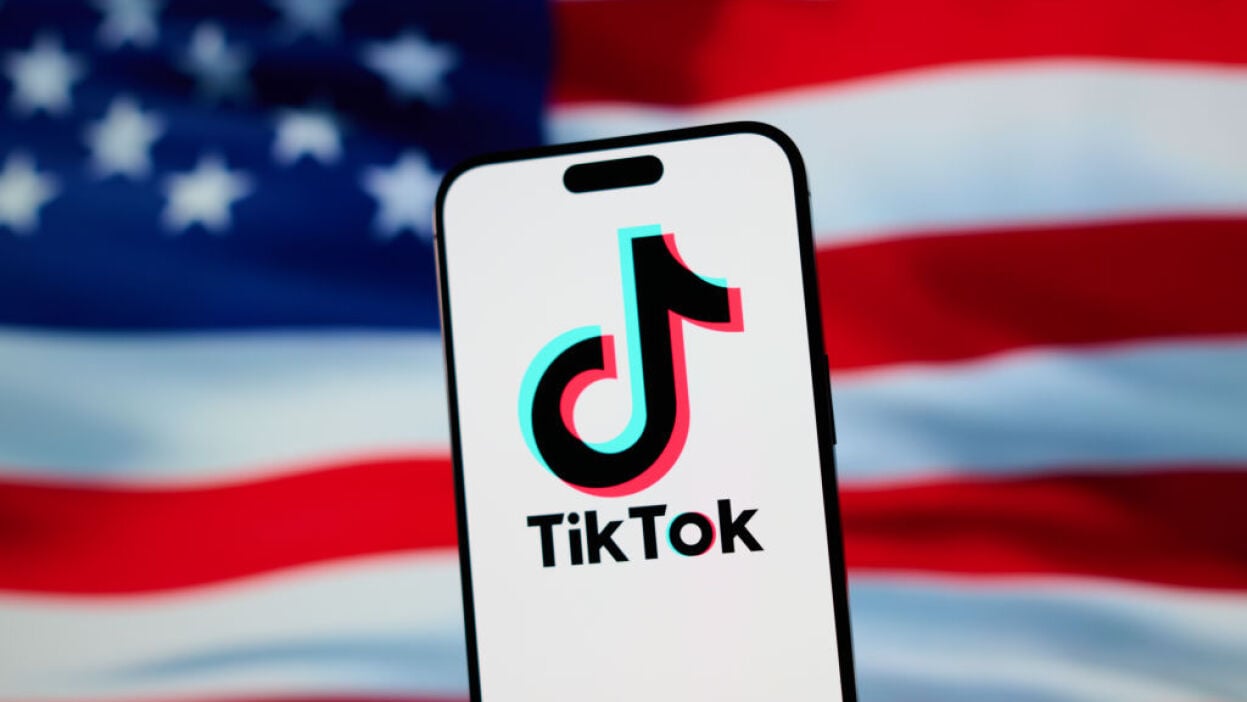 The TikTok app and logo are seen on a mobile device, the backdrop is the American flag. 