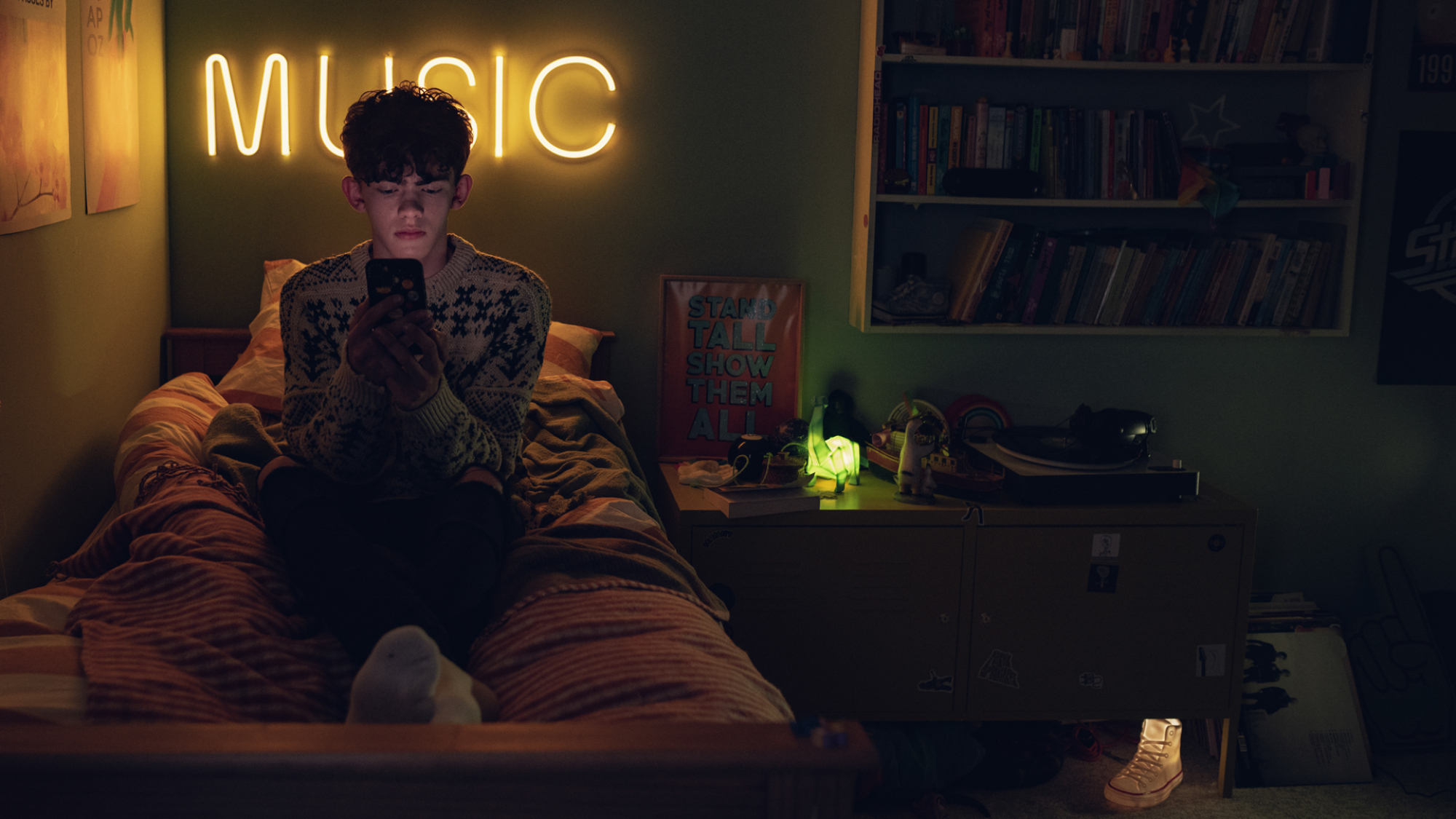 Joe Locke as Charlie in "Heartstopper" texting on his phone in his room, with a giant neon sign reading "Music."