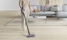 a person uses the Dyson v8 cordless stick vacuum on a brightly lit living room floor. behind her is a couch with a fluffy cat resting