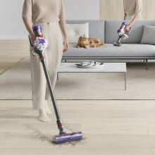 a person uses the Dyson v8 cordless stick vacuum on a brightly lit living room floor. behind her is a couch with a fluffy cat resting