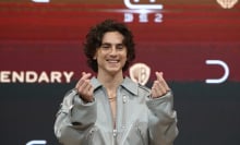 Timothée Chalamet smiling in a gray jumpsuit doing finger hearts.