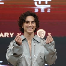 Timothée Chalamet smiling in a gray jumpsuit doing finger hearts.